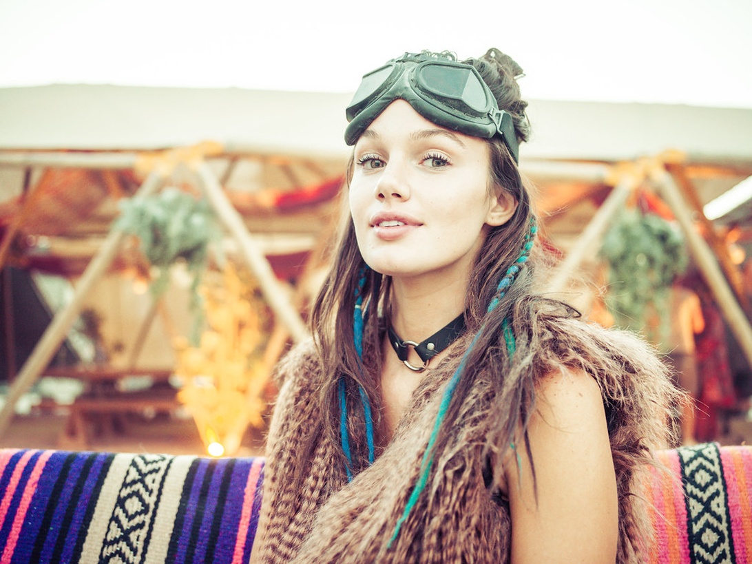 Kvinna som representerar en klassisk bohemisk stil med vintage-glasögon, färgstark fjästerväst och blågröna hårslingor, vilket skapar en festivalinspirerad boho-atmosfär.